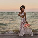 Stylish black bride with flower bouquet walking near ocean